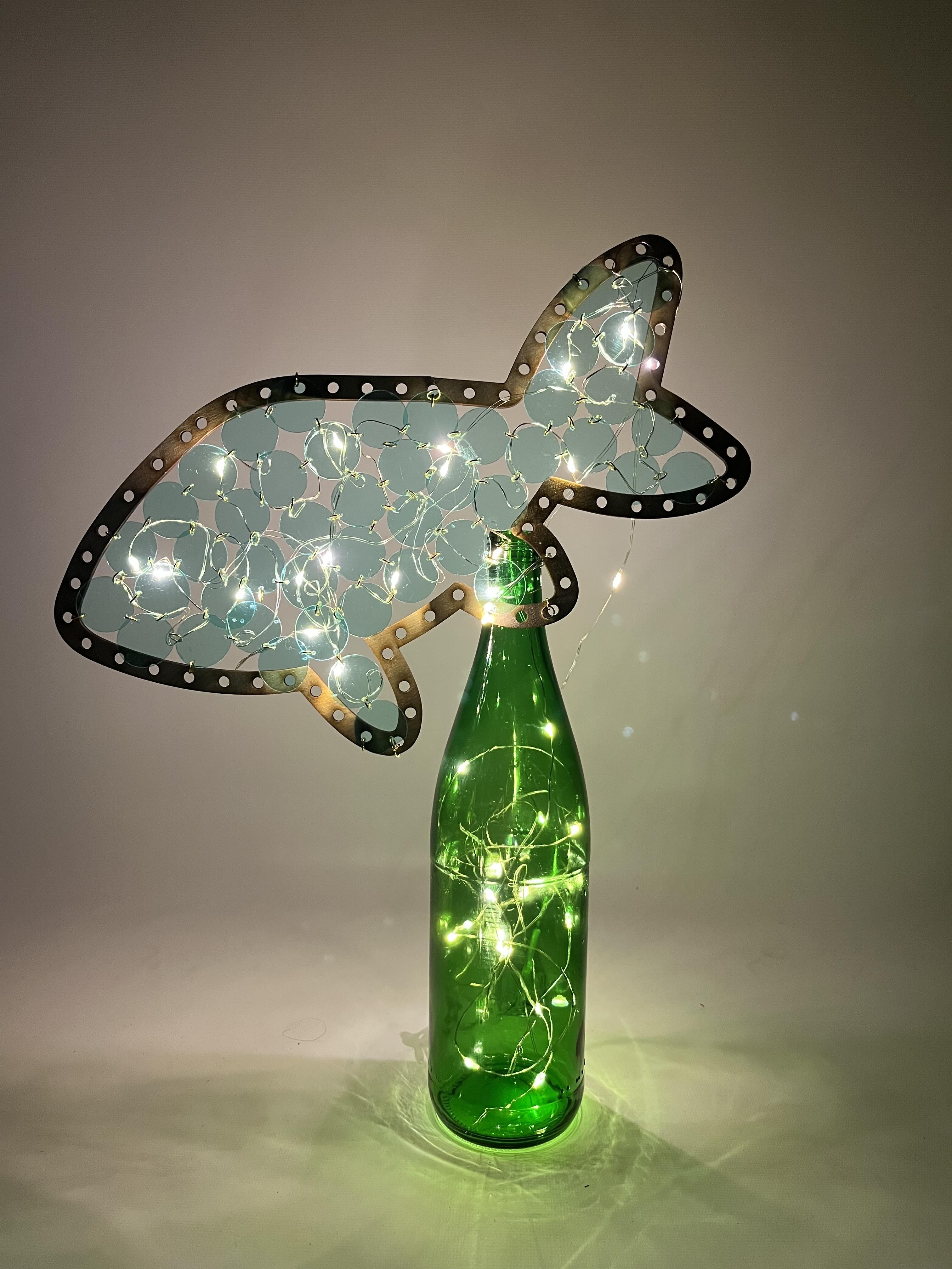 'Tilapia Fish Lamp Bottle', 2025. Upcycled green glass bottle, laser-cut plywood, woven PET plastic discs, LED fairy lights, and recycled wire